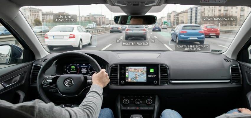 Modern autó belső tere a vezető szemszögéből, bemutatva az olyan alapfelszereltséggé vált technológiákat, mint a sávtartó, a vészfékasszisztens és az okostelefon-integráció.